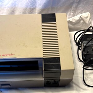 May include: A vintage Nintendo Entertainment System console in a beige and gray color scheme. The console has a cartridge slot, power and reset buttons, and the Nintendo logo. Includes a black power adapter.