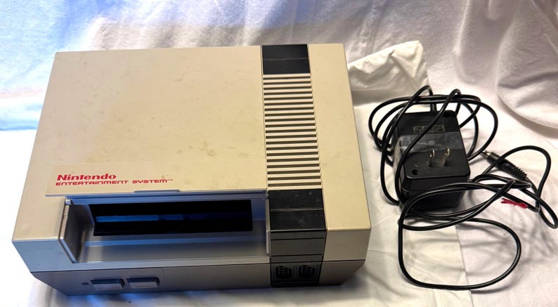 May include: A vintage Nintendo Entertainment System console in a beige and gray color scheme. The console has a cartridge slot, power and reset buttons, and the Nintendo logo. Includes a black power adapter.