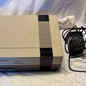 May include: A vintage Nintendo Entertainment System console in a beige and gray color scheme. The console has a black stripe down the center and the Nintendo logo is visible. A black power adapter and cables are next to the console.