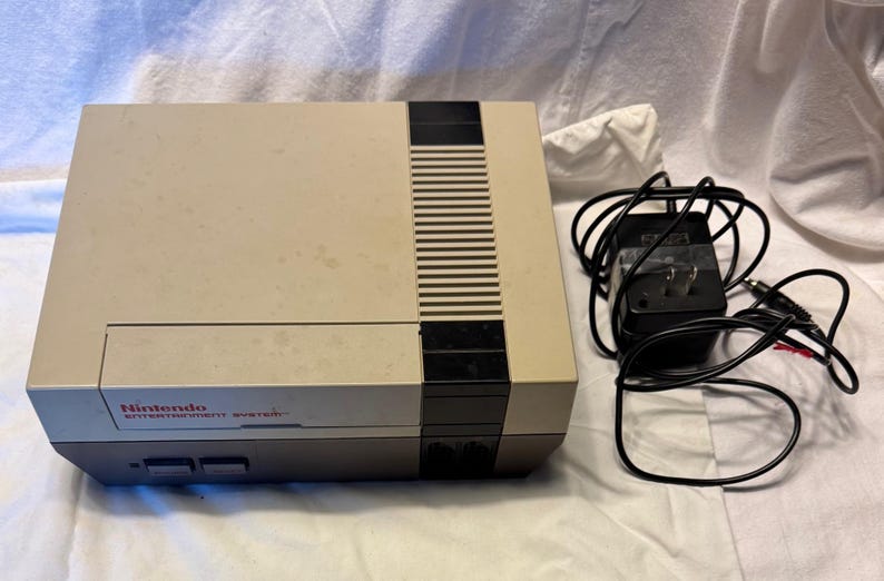 May include: A vintage Nintendo Entertainment System console in a beige and gray color scheme. The console has a black stripe down the center and the Nintendo logo is visible. A black power adapter and cables are next to the console.