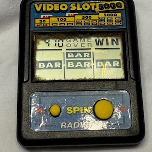 May include: A black handheld electronic game titled VIDEO SLOT 5000 with a blue control panel featuring two yellow buttons labeled SPIN. The screen displays the text GAME OVER, WIN, and a grid of BAR symbols with a numerical score of 410.