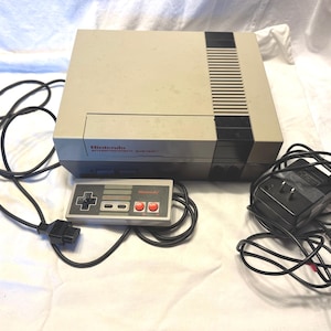 May include: A vintage Nintendo Entertainment System (NES) console with a gray and black color scheme. The console is accompanied by a controller and power adapter, all connected by black cords. The Nintendo logo is visible on the console and controller.