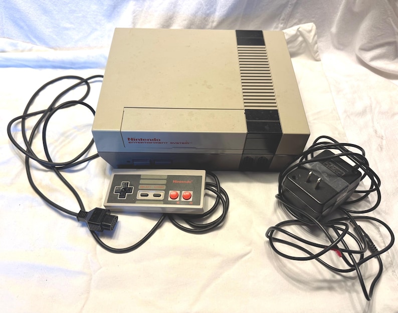 May include: A vintage Nintendo Entertainment System (NES) console with a gray and black color scheme. The console is accompanied by a controller and power adapter, all connected by black cords. The Nintendo logo is visible on the console and controller.