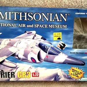 May include: A model kit box for a Harrier GR3 aircraft, featuring a detailed illustration of the plane in camouflage colors. The box is labeled "Smithsonian National Air and Space Museum" and includes the text "All in One Model Kit".