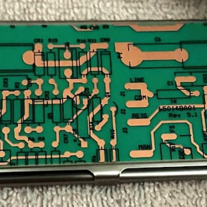 May include: A silver business card holder with a green circuit board design. The circuit board has copper-colored traces and the text "65014001 Rev 5.1". The holder is rectangular and has a hinged opening.