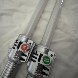 May include: Two toy lightsabers with clear blades and silver handles. One has a red button labeled "RED POWER", the other has a green button labeled "GREEN POWER". The handles have a ribbed design.