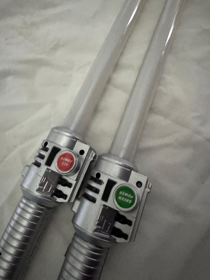 May include: Two toy lightsabers with clear blades and silver handles. One has a red button labeled "RED POWER", the other has a green button labeled "GREEN POWER". The handles have a ribbed design.