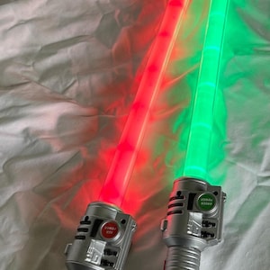 May include: Two toy light sabers with illuminated blades, one red and one green. The hilts are silver with buttons labeled "RED POWER" and "GREEN POWER". The blades are long and rectangular, and the toys are on a white surface.