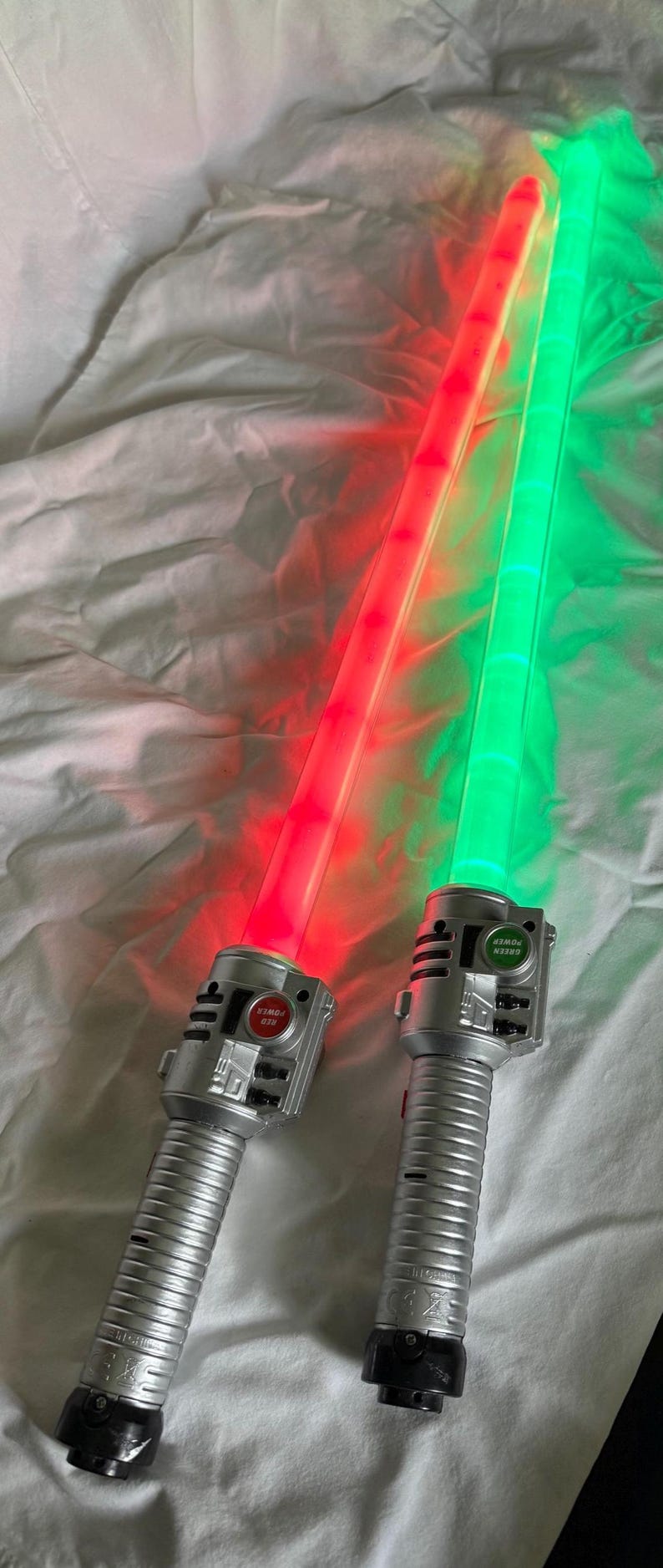 May include: Two toy light sabers with illuminated blades, one red and one green. The hilts are silver with buttons labeled "RED POWER" and "GREEN POWER". The blades are long and rectangular, and the toys are on a white surface.
