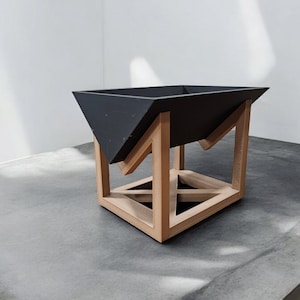 May include: A black geometric planter with a wooden stand. The planter is made of a smooth, matte material and has a square base. The stand is made of light brown wood and has a geometric design.