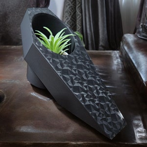 May include: A black geometric planter shaped like a coffin with a small green succulent plant inside. The planter is sitting on a dark brown surface with two black candles.