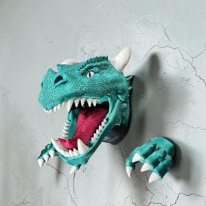 May include: A teal dragon head wall sculpture with an open mouth revealing a pink tongue and white teeth. The sculpture includes white horns and a teal clawed hand. The dragon is mounted on a textured gray wall.
