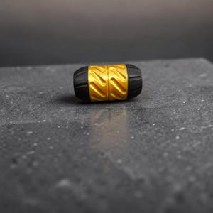 May include: A cylindrical bead featuring a central gold-colored section with diagonal grooves flanked by two black matte end caps. The bead has a textured surface and a symmetrical design, presenting a contrast between the metallic gold and black.