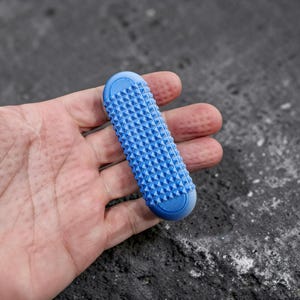 May include: A light blue, oblong-shaped sensory tool featuring a textured surface covered in small, uniform pyramid-shaped spikes. The ends of the object are rounded and feature a subtle, grooved, circular pattern on both the top and bottom edges.