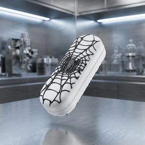 May include: An oblong white case featuring a raised black spider and web design on its top surface. The container has a smooth, rounded rectangular shape with a distinct black graphic pattern that covers the center of the lid.