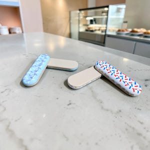 May include: Two rectangular, flat-topped objects with rounded ends. One features a light blue icing top with white sprinkles, while the other has a white icing top with red and blue sprinkles. Both have a plain, light beige underside.
