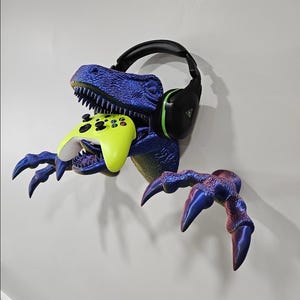 May include: A wall-mounted blue and purple iridescent dinosaur head sculpture with two extended claws. The dinosaur holds a bright neon yellow video game controller in its open mouth and wears black gaming headphones over its head.