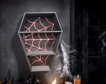 May include: A coffin-shaped, three-tiered shelf unit in dark gray, mounted on a black wall. Each shelf features a red spiderweb design against a black background, creating a Halloween theme. The shelves are empty, ready for display.