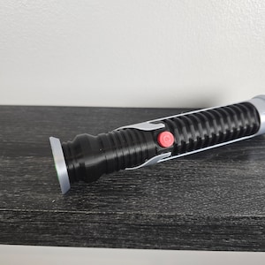 May include: A black lightsaber with a silver accent and a red button. The lightsaber is on a dark wood surface.
