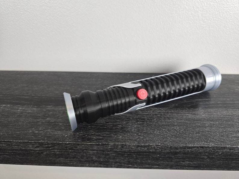May include: A black lightsaber with a silver accent and a red button. The lightsaber is on a dark wood surface.