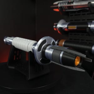 Peut inclure: Un sabre laser argent&eacute; et noir avec une poign&eacute;e grise, expos&eacute; sur un support noir. Le sabre laser a une garde circulaire et une lueur orange &agrave; l'extr&eacute;mit&eacute;. D'autres sabres laser sont visibles en arri&egrave;re-plan.