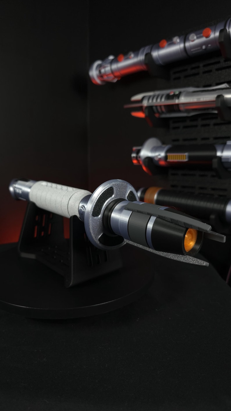 Peut inclure: Un sabre laser argent&eacute; et noir avec une poign&eacute;e grise, expos&eacute; sur un support noir. Le sabre laser a une garde circulaire et une lueur orange &agrave; l'extr&eacute;mit&eacute;. D'autres sabres laser sont visibles en arri&egrave;re-plan.