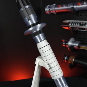 Peut inclure: Image d&eacute;taill&eacute;e d'un sabre laser avec un manche argent&eacute; et blanc et un &eacute;metteur noir. Le sabre laser est pr&eacute;sent&eacute; sur un support blanc, avec d'autres sabres laser en arri&egrave;re-plan. L'esth&eacute;tique g&eacute;n&eacute;rale est futuriste et de science-fiction.