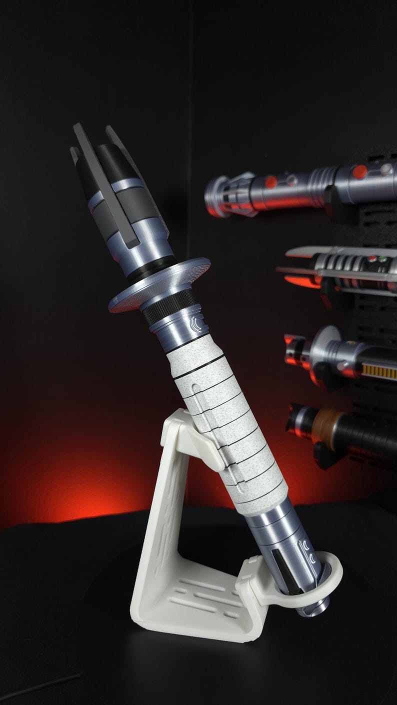 Peut inclure: Image d&eacute;taill&eacute;e d'un sabre laser avec un manche argent&eacute; et blanc et un &eacute;metteur noir. Le sabre laser est pr&eacute;sent&eacute; sur un support blanc, avec d'autres sabres laser en arri&egrave;re-plan. L'esth&eacute;tique g&eacute;n&eacute;rale est futuriste et de science-fiction.