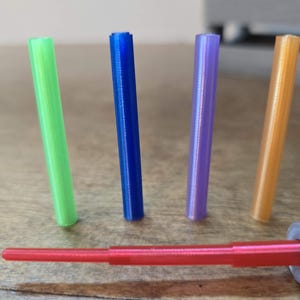 Peut inclure: Une collection d'objets cylindriques color&eacute;s. Quatre cylindres verticaux verts, bleus, violets et oranges sont pr&eacute;sent&eacute;s. Une tige rouge &agrave; plusieurs niveaux avec un raccord argent&eacute; et dor&eacute; est en bas. Les objets sont sur une surface en bois.