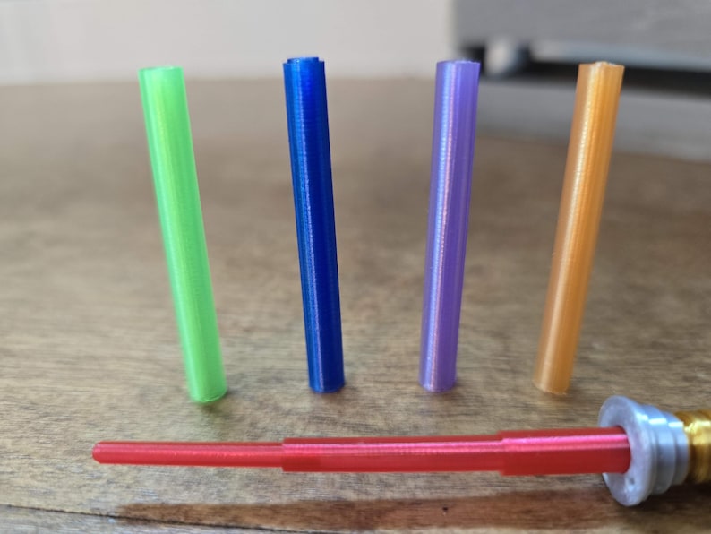 Peut inclure: Une collection d'objets cylindriques color&eacute;s. Quatre cylindres verticaux verts, bleus, violets et oranges sont pr&eacute;sent&eacute;s. Une tige rouge &agrave; plusieurs niveaux avec un raccord argent&eacute; et dor&eacute; est en bas. Les objets sont sur une surface en bois.