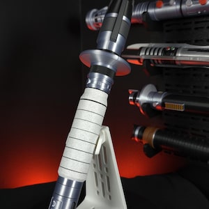 Peut inclure: Un sabre laser argent&eacute; et noir, avec une poign&eacute;e blanche et une garde circulaire, est expos&eacute; sur un support blanc. D'autres sabres laser sont visibles en arri&egrave;re-plan, mont&eacute;s sur une surface noire. Le sabre laser est une r&eacute;plique.