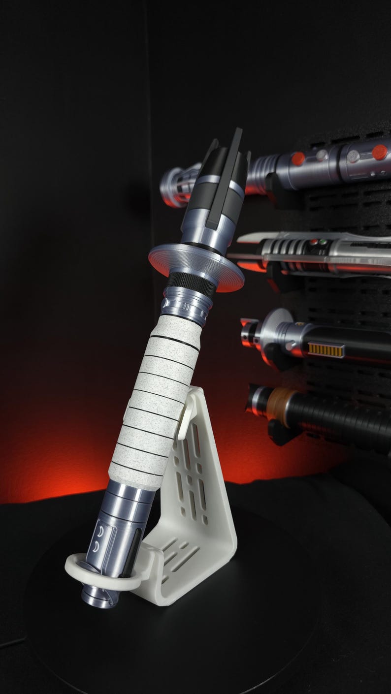 Peut inclure: Un sabre laser argent&eacute; et noir, avec une poign&eacute;e blanche et une garde circulaire, est expos&eacute; sur un support blanc. D'autres sabres laser sont visibles en arri&egrave;re-plan, mont&eacute;s sur une surface noire. Le sabre laser est une r&eacute;plique.