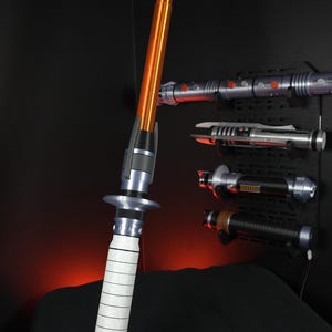 Peut inclure: Un sabre laser orange et argent&eacute; avec une poign&eacute;e blanche est pr&eacute;sent&eacute;. Plusieurs autres sabres laser sont mont&eacute;s sur un mur noir en arri&egrave;re-plan. Les sabres laser sont un article populaire dans le genre de la science-fiction.