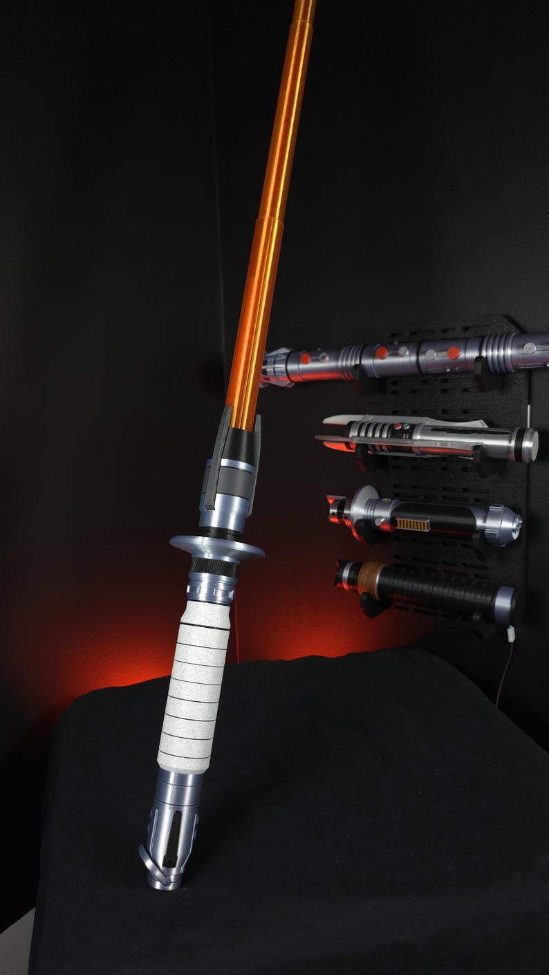 Peut inclure: Un sabre laser orange et argent&eacute; avec une poign&eacute;e blanche est pr&eacute;sent&eacute;. Plusieurs autres sabres laser sont mont&eacute;s sur un mur noir en arri&egrave;re-plan. Les sabres laser sont un article populaire dans le genre de la science-fiction.