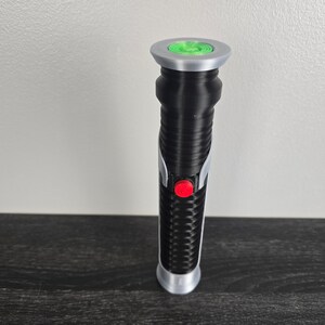 May include: A black lightsaber toy with a silver handle and a green light at the end. The lightsaber has a red button on the side.