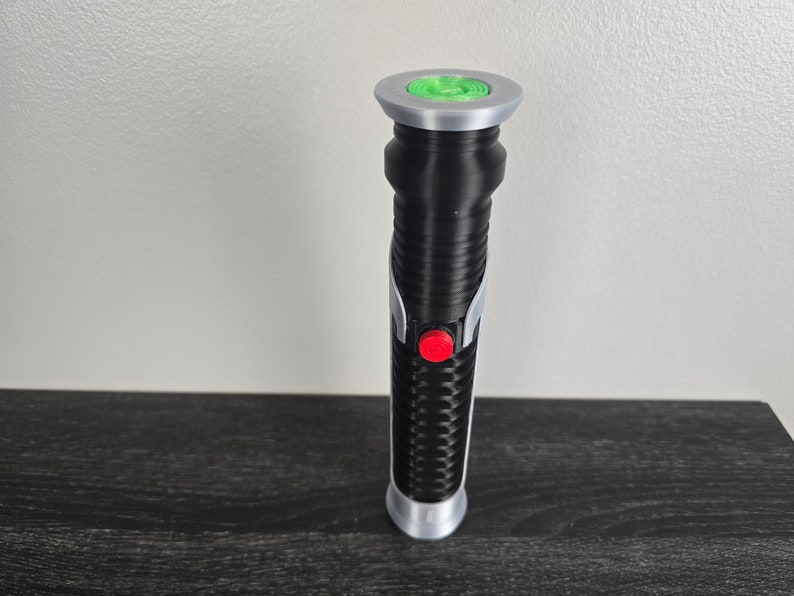 May include: A black lightsaber toy with a silver handle and a green light at the end. The lightsaber has a red button on the side.