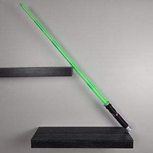 May include: A green lightsaber toy with a black handle rests on a black shelf. The lightsaber is angled diagonally, pointing towards the right side of the image.
