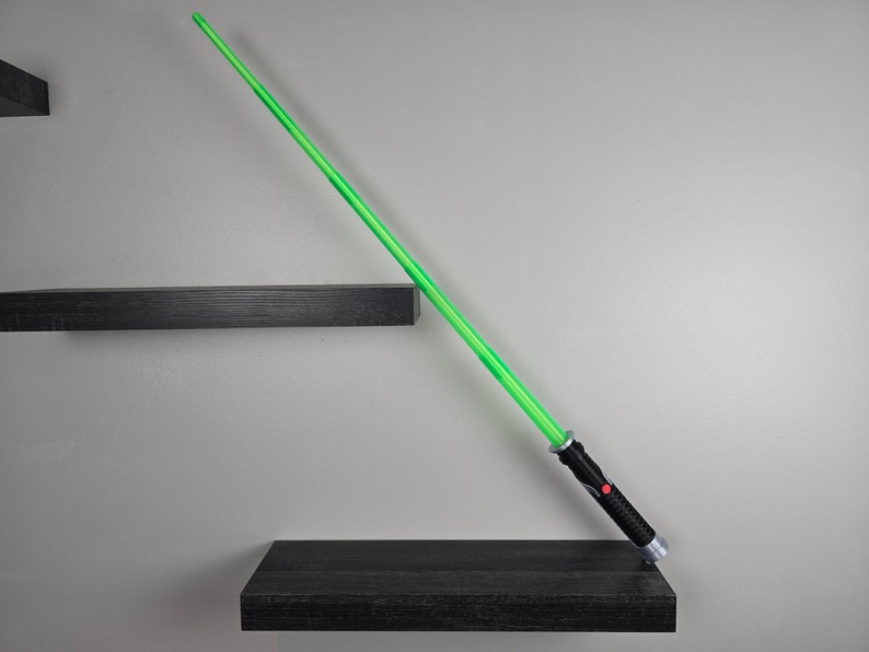 May include: A green lightsaber toy with a black handle rests on a black shelf. The lightsaber is angled diagonally, pointing towards the right side of the image.