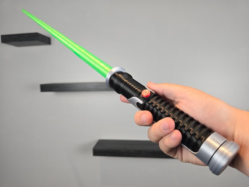 May include: A black and silver lightsaber toy with a green blade. The lightsaber has a red button on the side.
