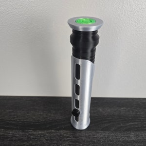 May include: A silver lightsaber hilt with a black grip and a green button on the top.