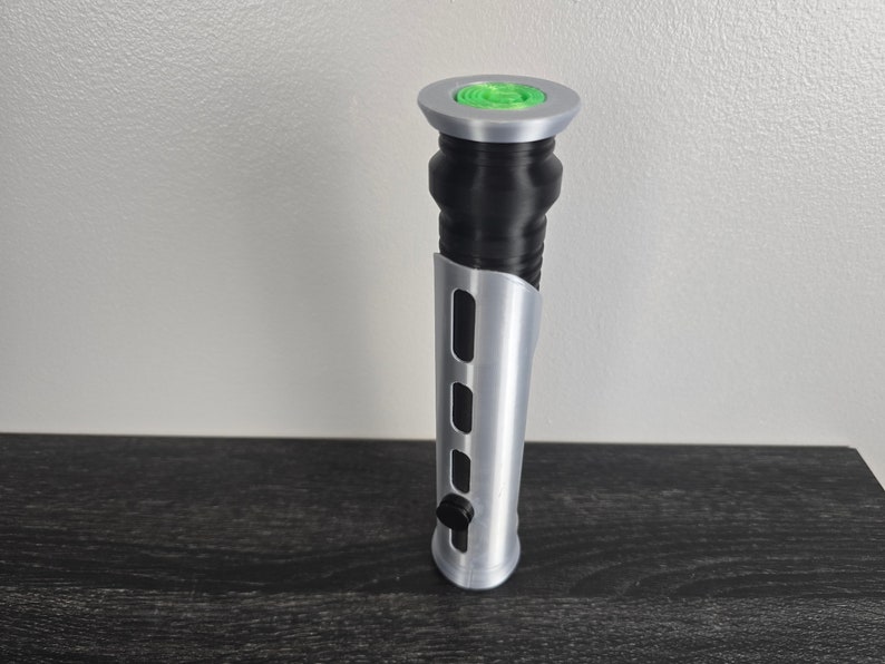May include: A silver lightsaber hilt with a black grip and a green button on the top.