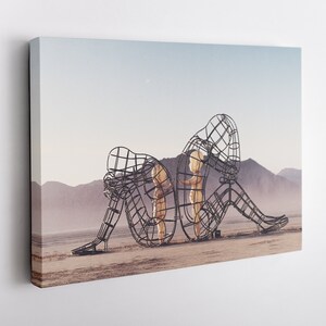 May include: A large metal sculpture of two figures, one sitting and the other standing, in a desert landscape. The figures are made of a wire mesh and appear to be embracing.