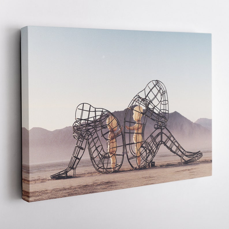 May include: A large metal sculpture of two figures, one sitting and the other standing, in a desert landscape. The figures are made of a wire mesh and appear to be embracing.