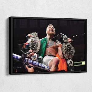 May include: A mixed martial arts fighter wearing a white and black uniform holds two championship belts, one in each hand. He is smiling and looking up. The belts are silver and black with gold accents. The fighter is standing in a ring with a crowd in the background.