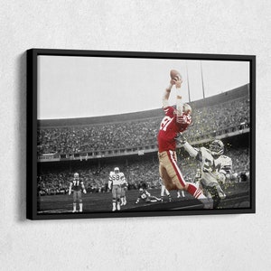 May include: A black and white photograph of a football player in a red and gold uniform jumping to catch a football. The player is surrounded by other players and a large crowd of spectators.