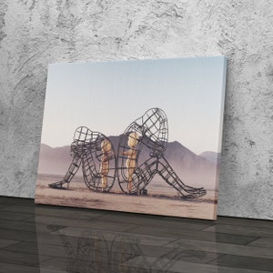 May include: A black metal sculpture of three figures in a desert landscape. The figures are made of wire mesh and appear to be embracing each other. The sculpture is set against a backdrop of a blue sky and distant mountains.