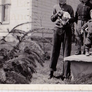 May include: A black and white photo of four people standing outside. Two children are wearing hats, and one is holding a small white animal. The adults are wearing coats and hats. The photo is taken in front of a building with a wooden door.