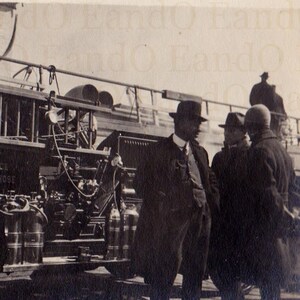 May include: A vintage fire engine with a ladder and hoses. The engine is parked in front of a building and several people are standing around it. The engine has the words "Angle G Hose" on the side.