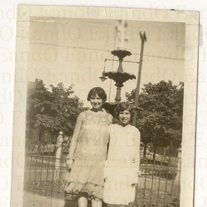 May include: Two young women in dresses stand in front of a fountain in a park. The woman on the left is wearing a white dress with lace detailing. The woman on the right is wearing a white dress with a simple design. Both women are smiling.