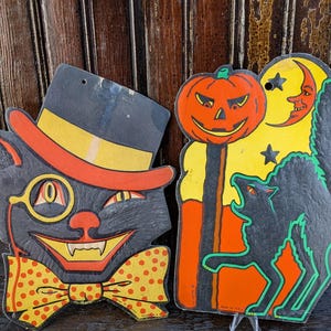 May include: Vintage Halloween decorations. One is a black cat wearing a top hat and monocle, the other a pumpkin with a wolf and moon. The cat has a yellow bow tie with red spots. The pumpkin is orange with a black face.
